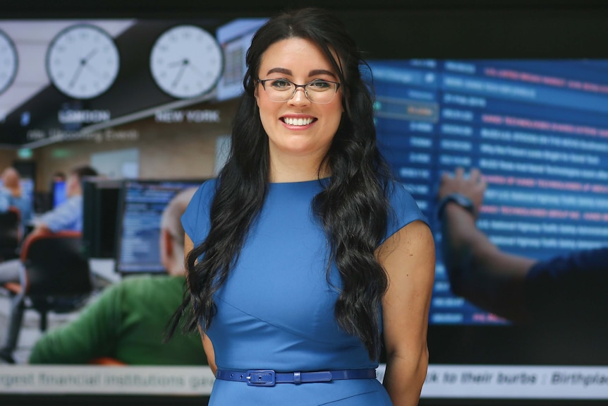 Jana Bowden has long wavy black hair, glasses and a blue dress and smiles in front of images of an office