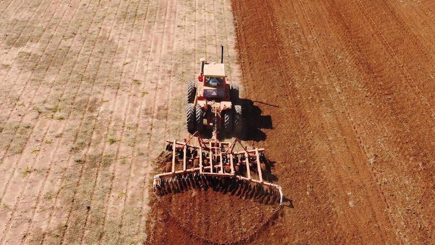 An overhead shot taken by a drone of a tractor in a paddock. Half the paddock is soil, the other half is weed