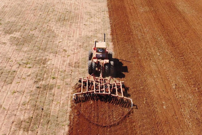 An overhead shot taken by a drone of a tractor in a paddock. Half the paddock is soil, the other half is weed