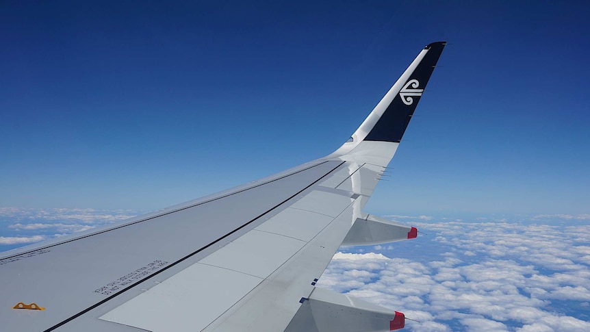 You look out over a plane wing flying over patchy clouds scattered across a blue sky with the Air New Zealand logo on the tip.