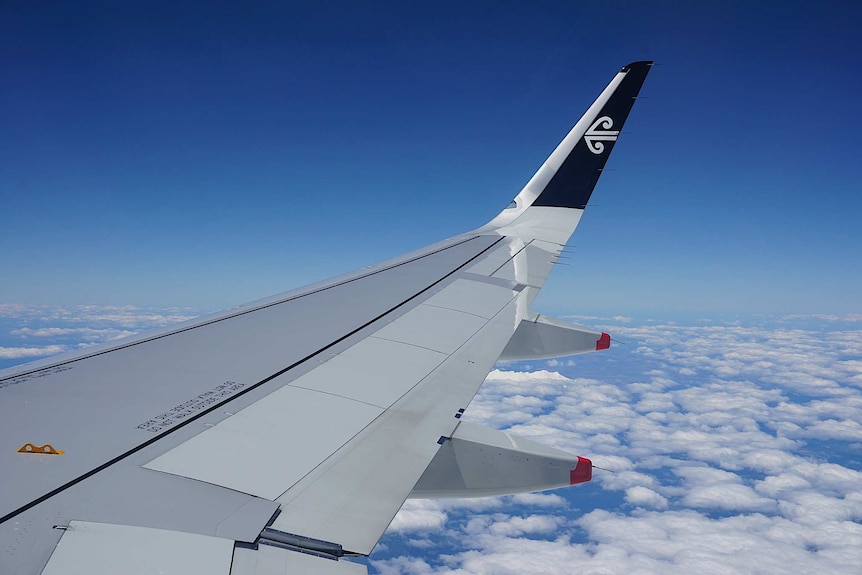 You look out over a plane wing flying over patchy clouds scattered across a blue sky with the Air New Zealand logo on the tip.