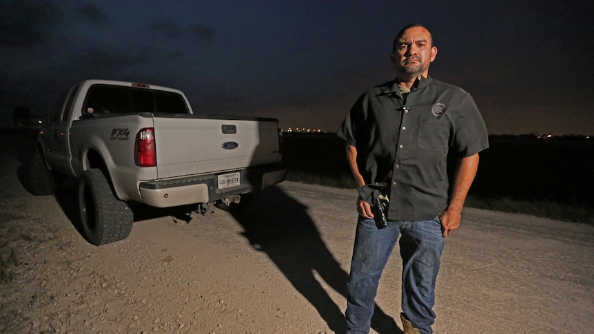 A border patrol agent stands next to his truck, holding a handgun.