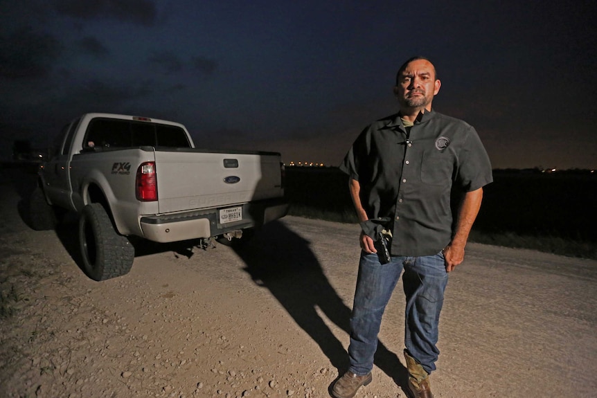 A border patrol agent stands next to his truck, holding a handgun.