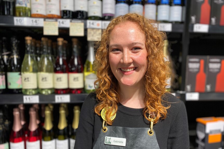 Gabriella Rush, a young woman with orange hair, smiles happily in front of a display of wine.