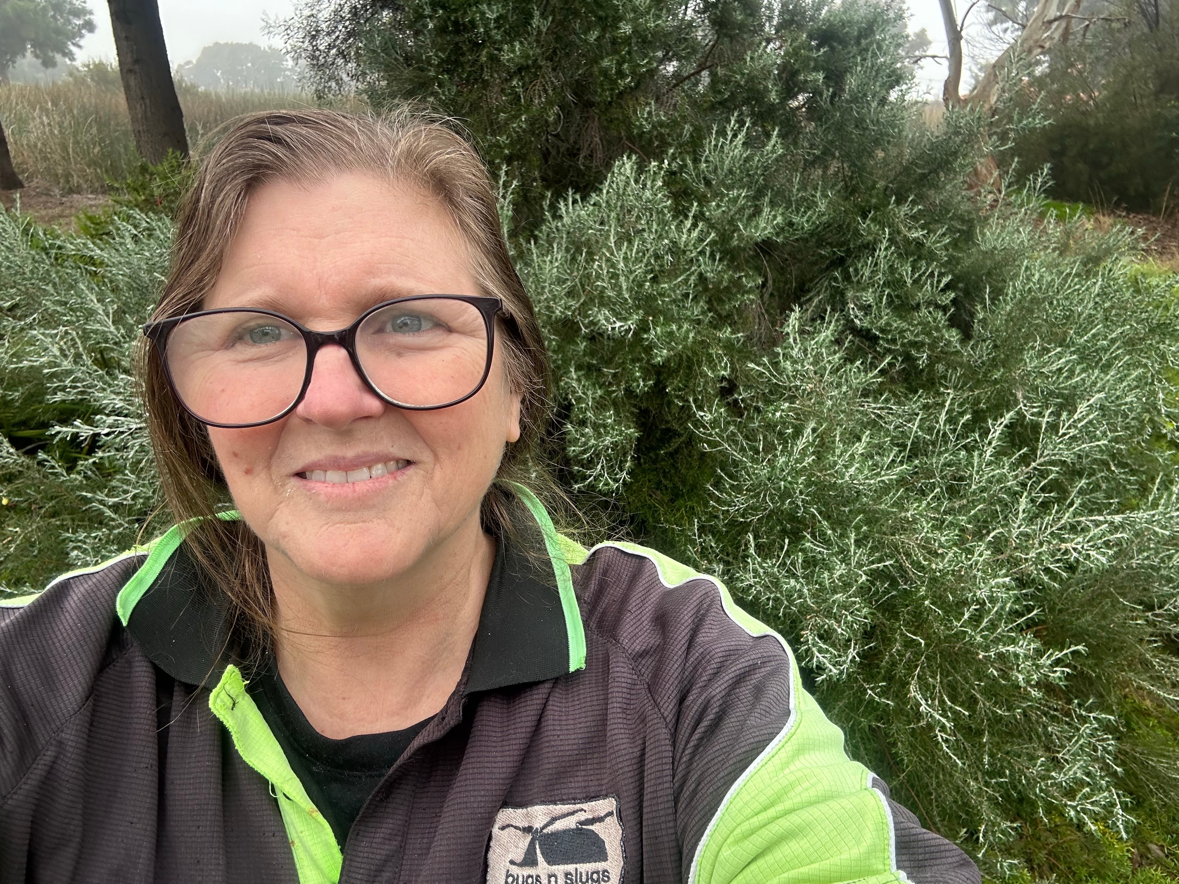 A woman wearing glasses smiling at the camera with a green bush in the background