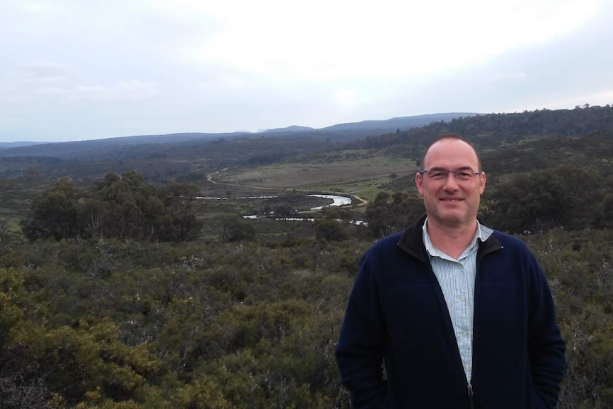 Wide shot of a man facing the camera with a serpentine river in the background. Man has glasses and is smiling