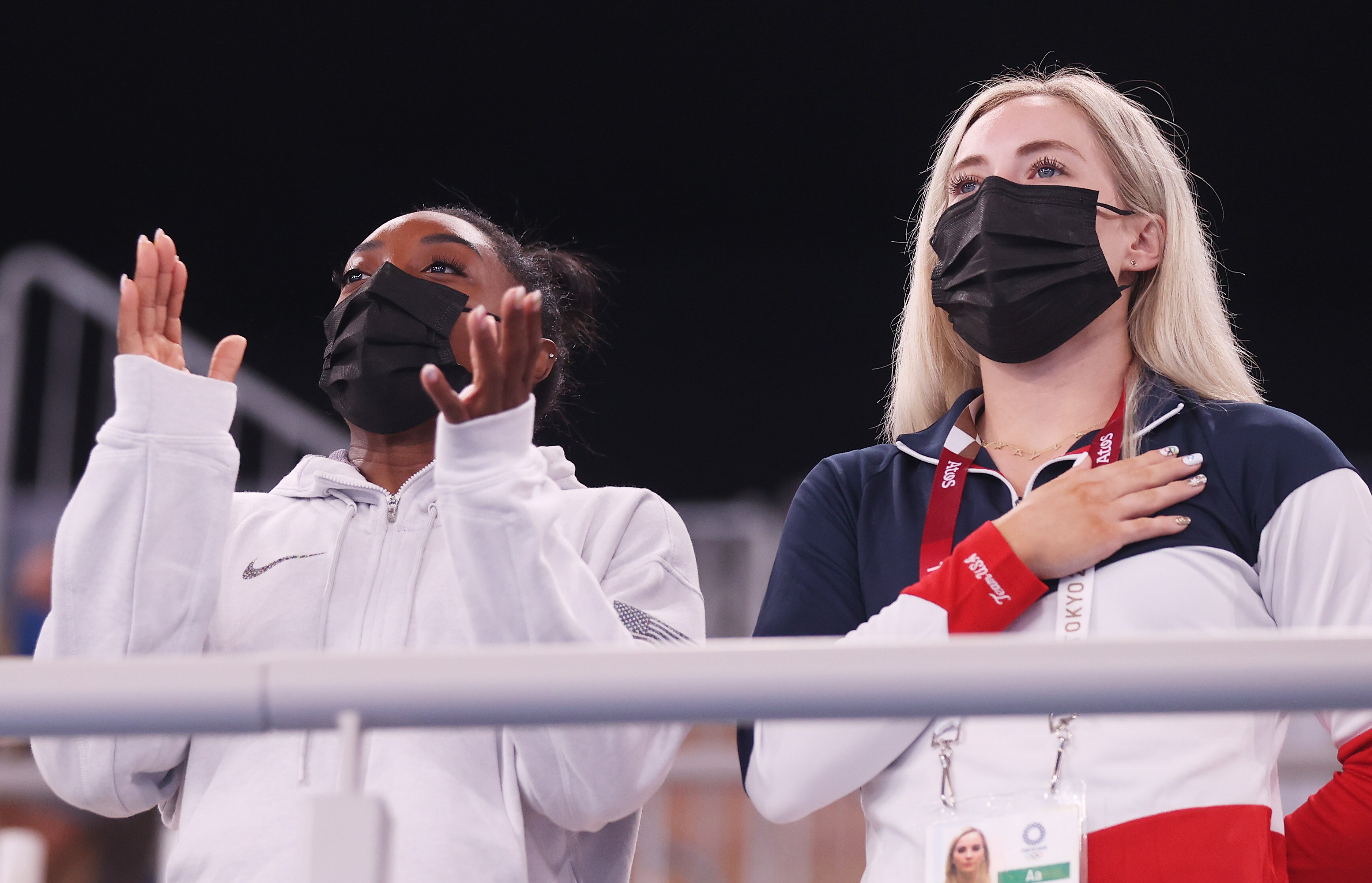 Two women wear masks at a sporting event, one claps the other has her hand on her heart.