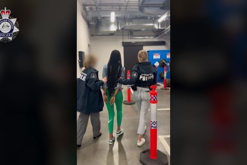 Two female police officers escort a woman in a car park with her hands handcuffed behind her.