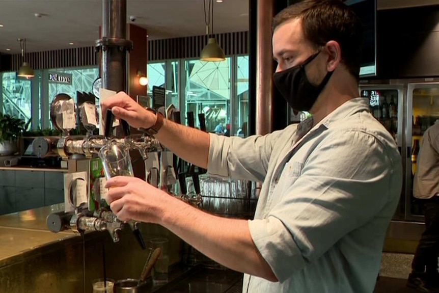 Luke Murphy pours a beer from the tap while wearing a black face mask.