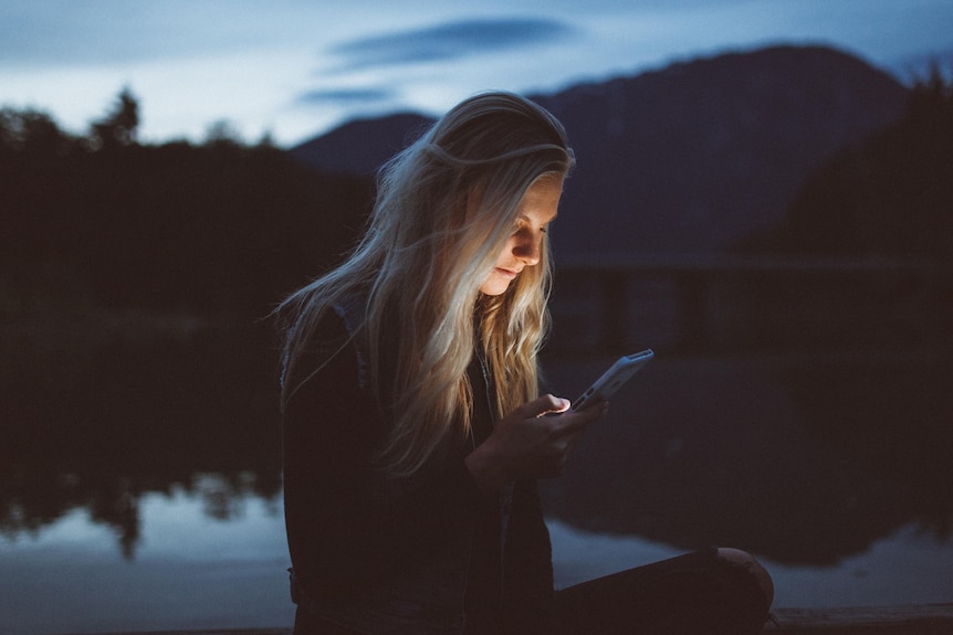 A generic photo of a woman by a lake looking at her phone at dusk, date unknown.