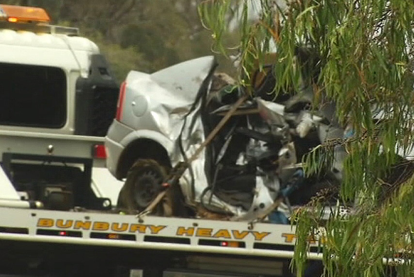 A smashed car sits on the bed of a tow truck, with a tree in the foreground.