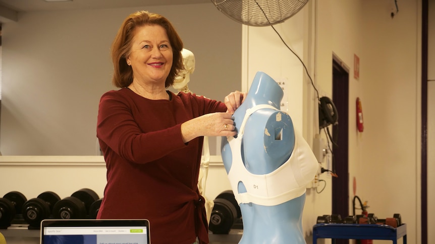 Deirdre McGhee fits a white sports bra on a blue mannequin while smiling and wearing a maroon jumper.