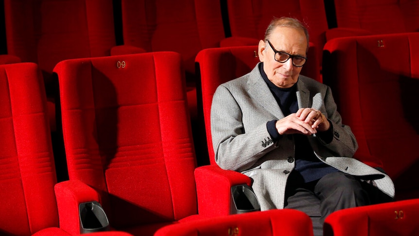 A man in a jacket sits in a theatre with red seats.