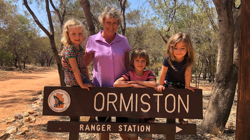 Ewart with three children in front of Ormiston sign with bush in background.