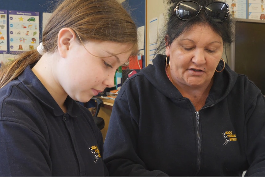 A young girl sits on the left in a navy polo, a middle aged woman in the same polo sits to her right both talking, looking down