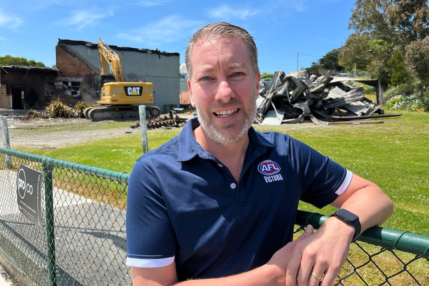 A man in a blue sports shirt stands beside a fence with a burnt-out building in the background.