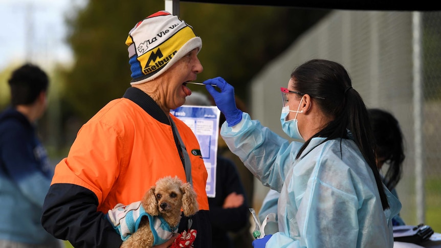 A man in an orange shirt and colourful hat holding a dog gets tested for COVID-19.