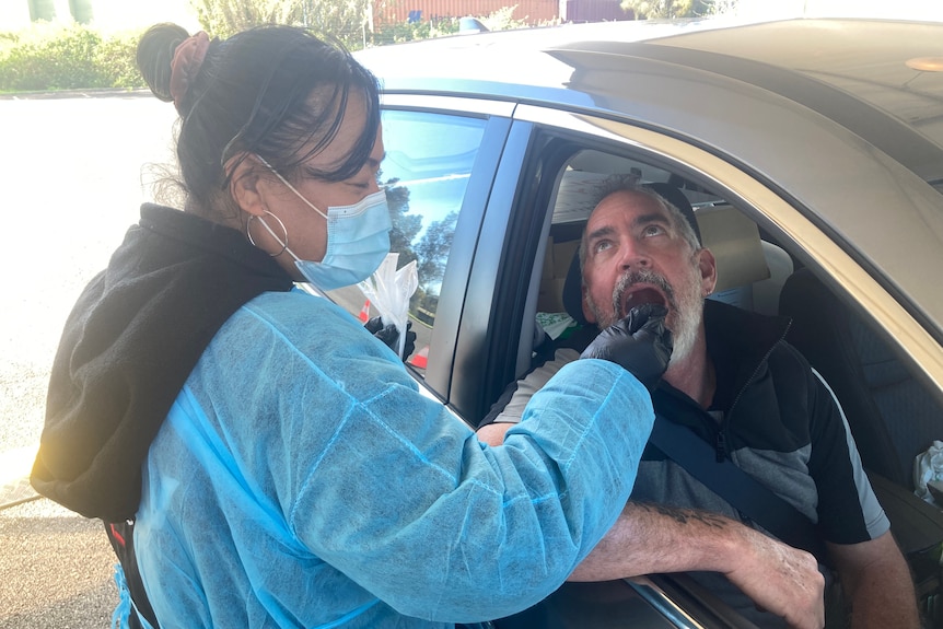 A woman in mask and blue scrubs takes a swab from the mouth of a man in a car.