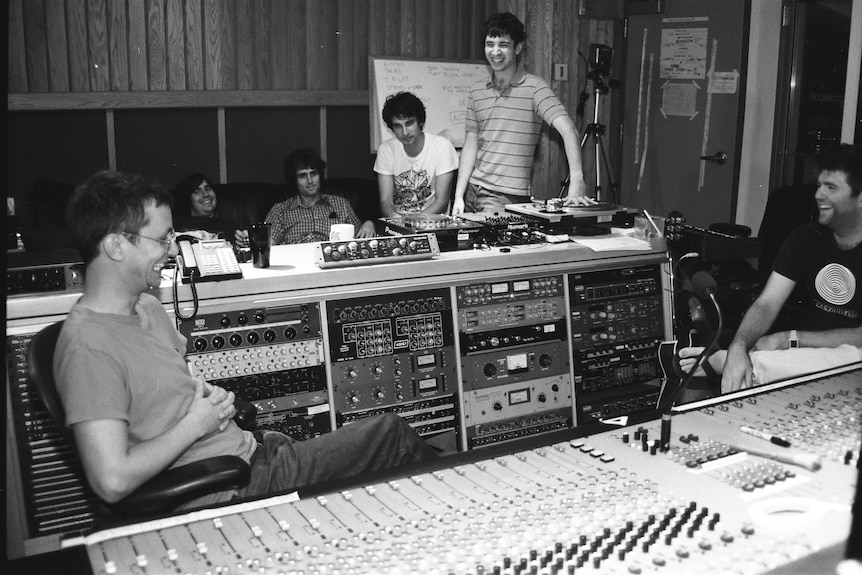 A B&W photograph of six young men sitting in a recording studio, laughing. Two are behind the studio desk.