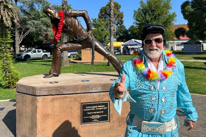 man in bright blue suit, black wig and flower lei dances in front of a bronze statue