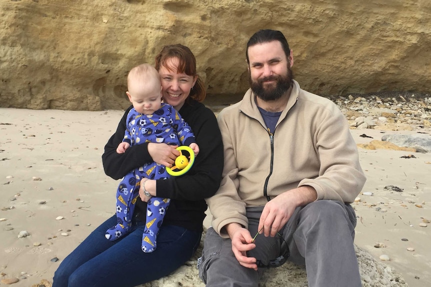 Laura Henning, Chris Hombsch with their baby Victor sitting on a beach in front of a sandstone cliff