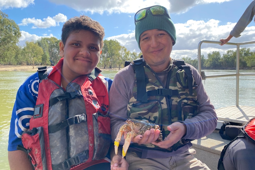 Two indigenous men smile, they wear life jackets sitting on a boat, they hold a medium sized crayfish with white claws. 