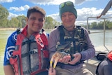 Two indigenous men smile, they wear life jackets sitting on a boat, they hold a medium sized crayfish with white claws. 