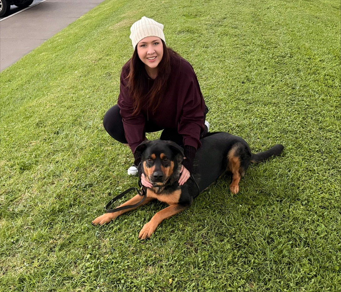 A smiling woman squats down behind a big dog lying down on the grass, giving it a hug.