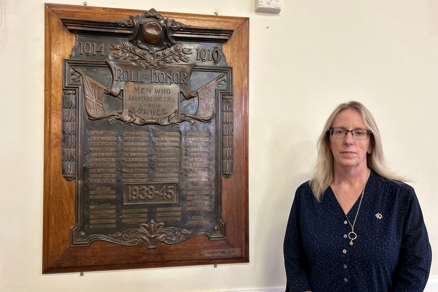 A woman stands next to a memorial plaque hanging from a wall in a town hall.