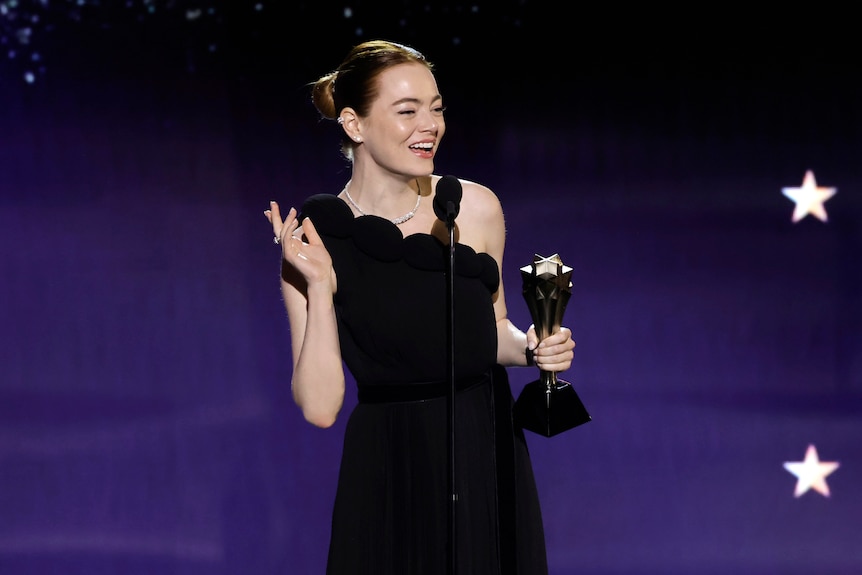 Emma, in a black gown, gestures and smiles on stage as she accepts her award