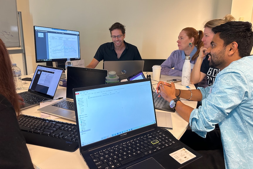 A group of people sit around computers looking at a larger screen.