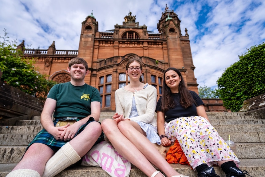 A young man wearing a kilt and two young women sitting on steps outdoors and smiling at the camera.