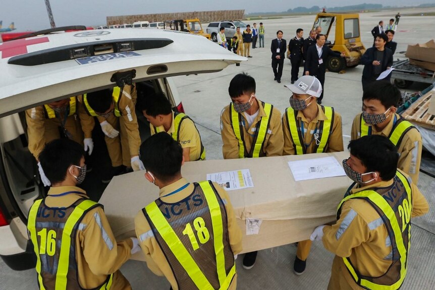 A group of men in hi-vis vests carry a coffin wrapped in beige fabric and load it into a white ambulance.