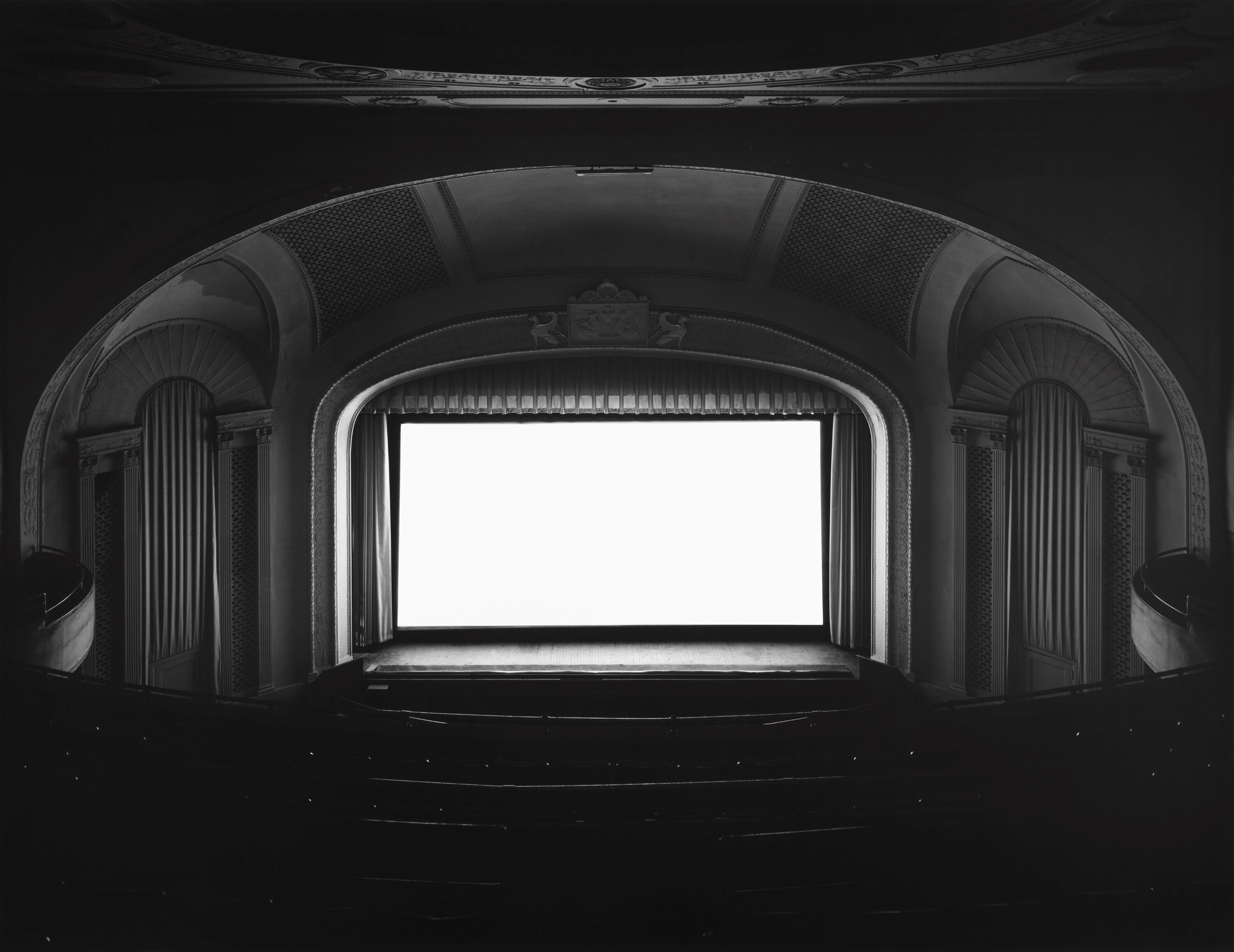 A black and white photograph of the interior of a large cinema, the screen entirely white