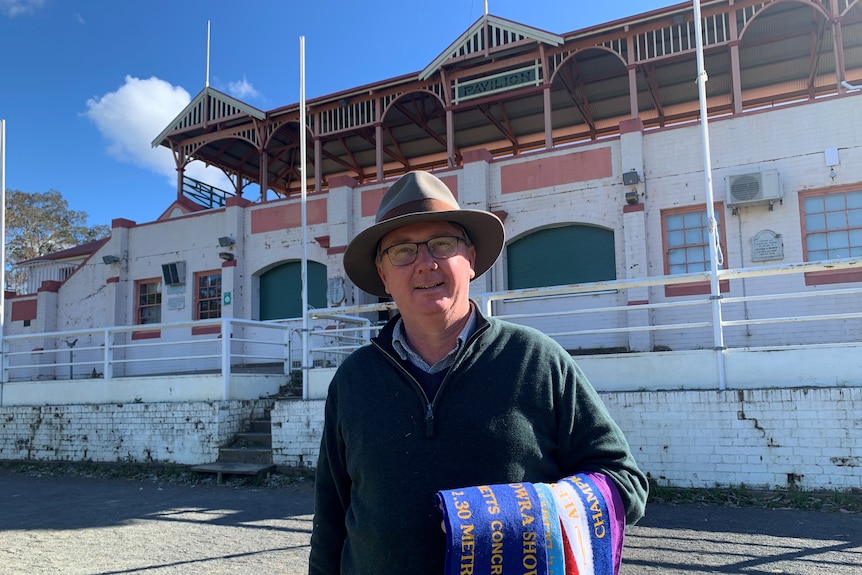 A man stands with ribbons on his arm in front of an old show pavillon.