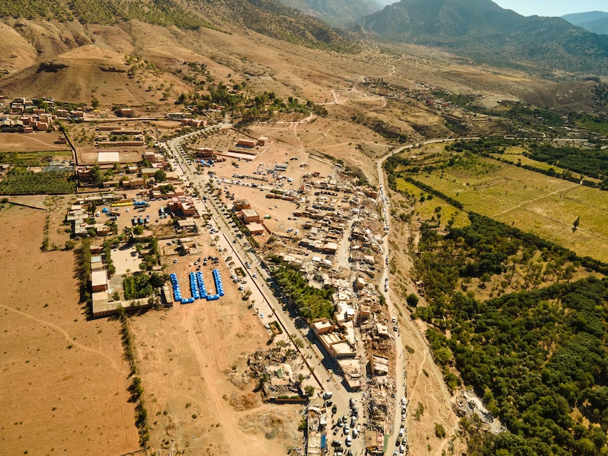 A town is seen from above, several buildings are destroyed, blue relief tents visible