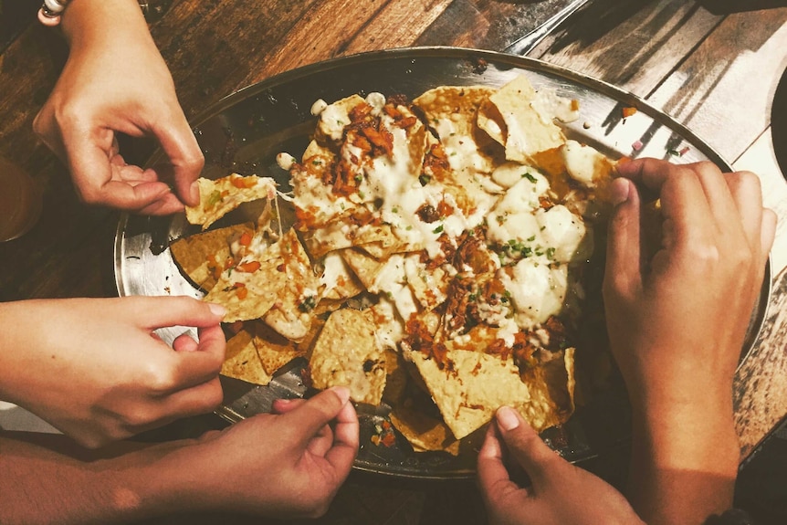 Hands reaching for nacho chips on a black oval plate.