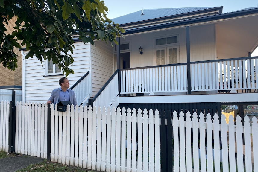 A man stands behind a fence looking back at a Queenslander home.