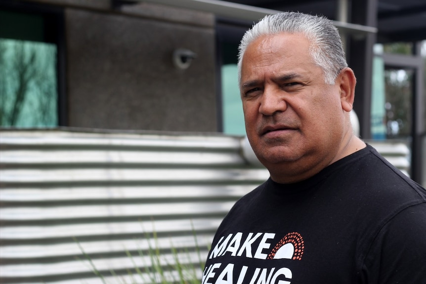 An Indigenous man with short, salt-and-pepper hair stands in front of a building, looking solemn.