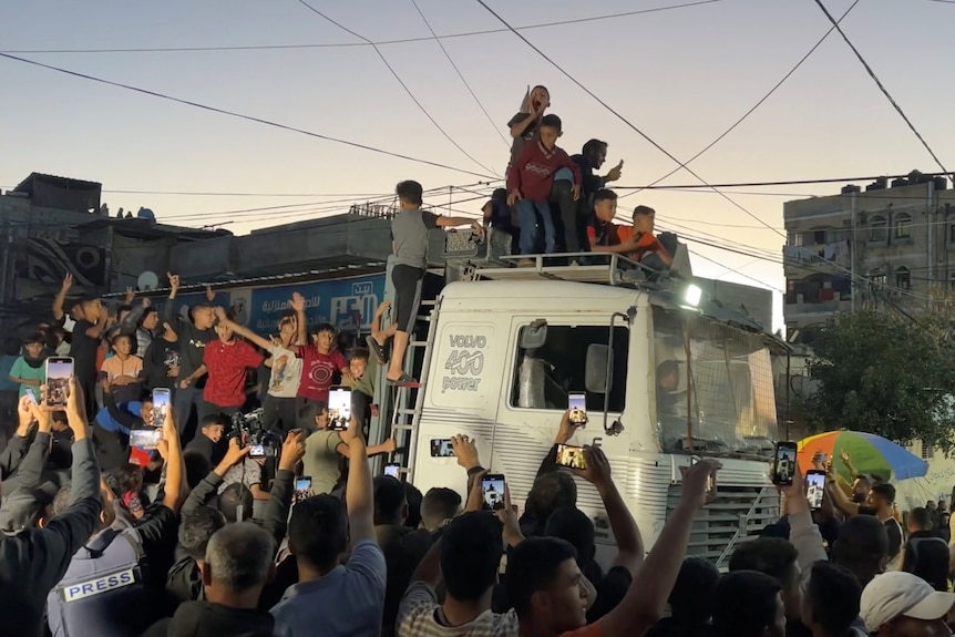 People climb on a truck, shouting and celebrating, while a people around the truck hold phones and film