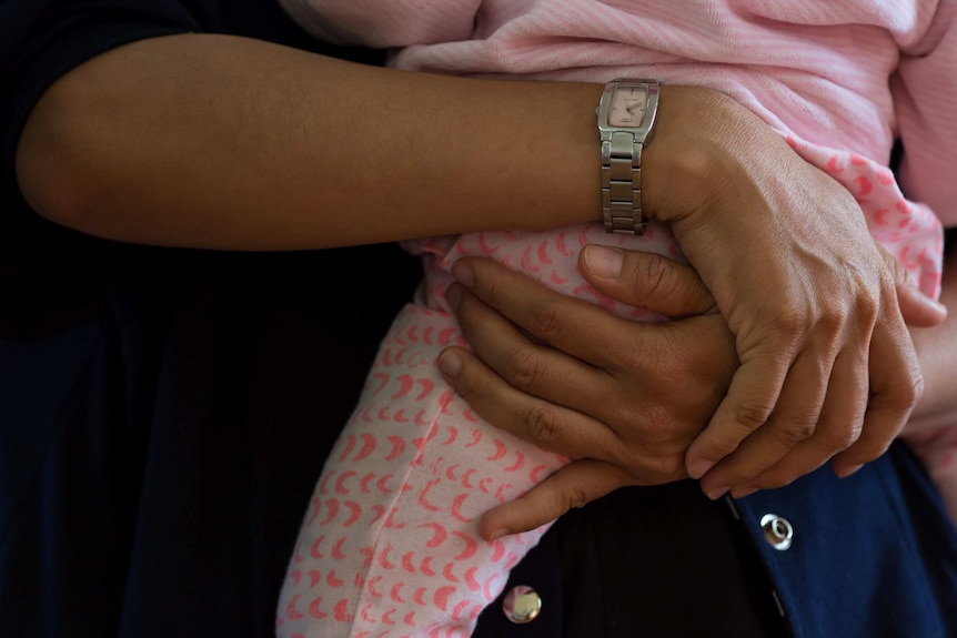 A close up of Layla's hands, with a watch on her right wrist, has she holds Tia, who is wearing a pink playsuit.
