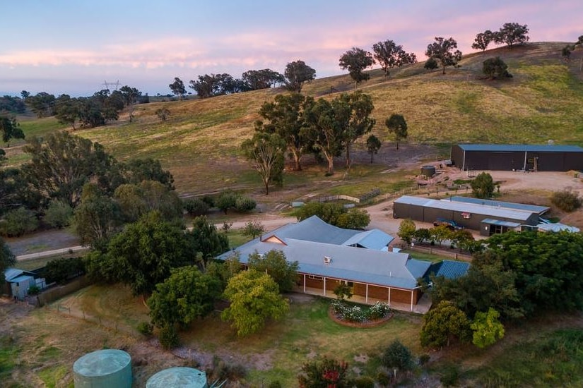 A large rural property with water tanks is nestled in a slope of partly cleared bushland.