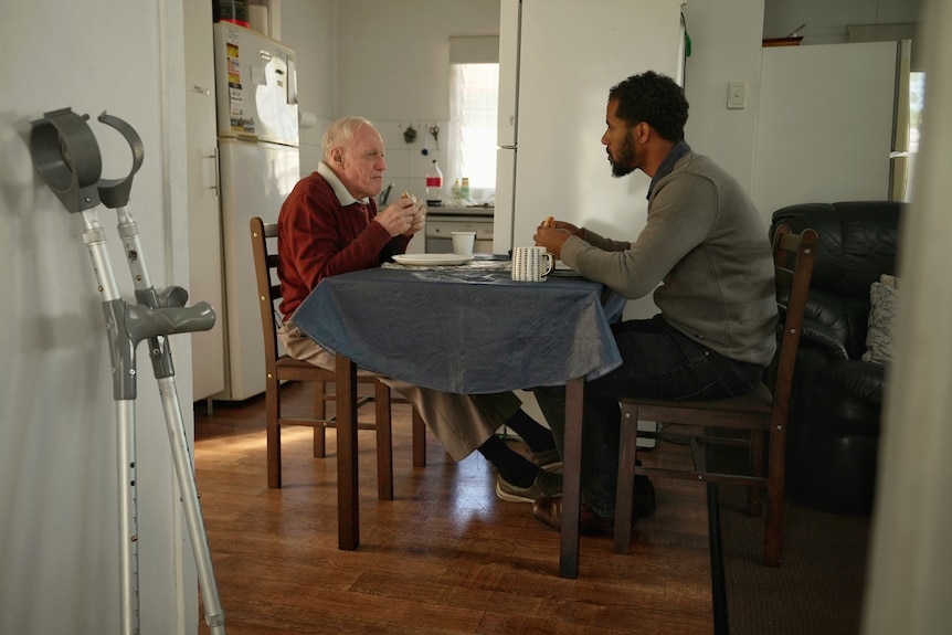 Two men sit at a dining table, eating. A pair of crutches is in the foreground.