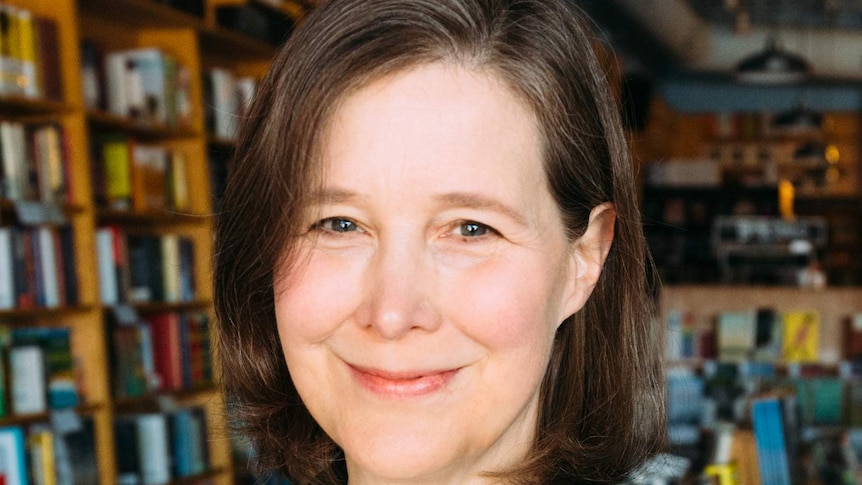 A woman stands with a book shop in the background, smiling, wearing a white shirt and black blazer