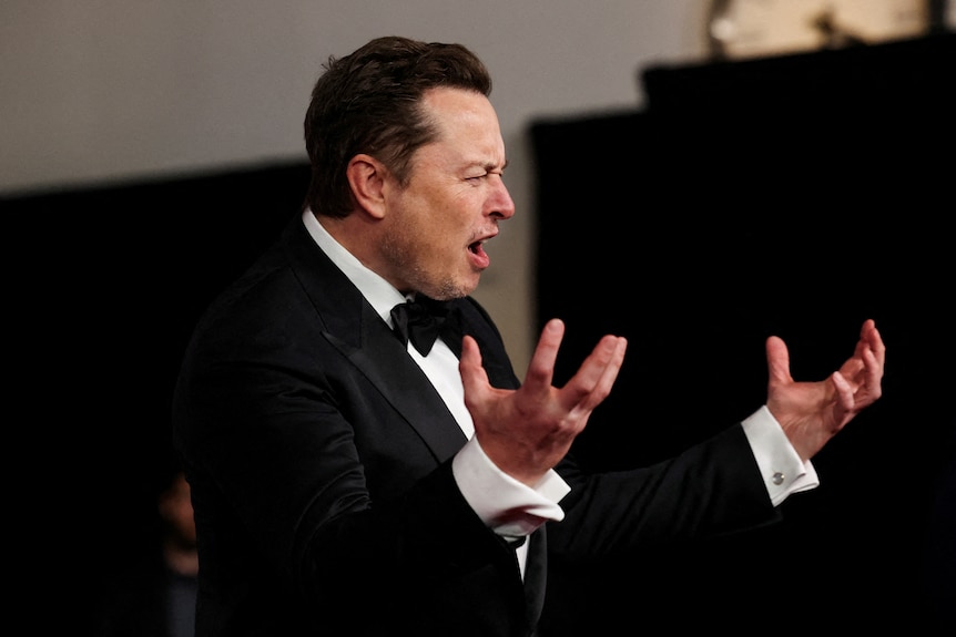 Elon Musk holds both hands out in front of him while contorting his mouth into a horrified expression