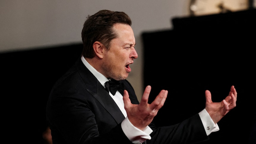 Elon Musk holds both hands out in front of him while contorting his mouth into a horrified expression