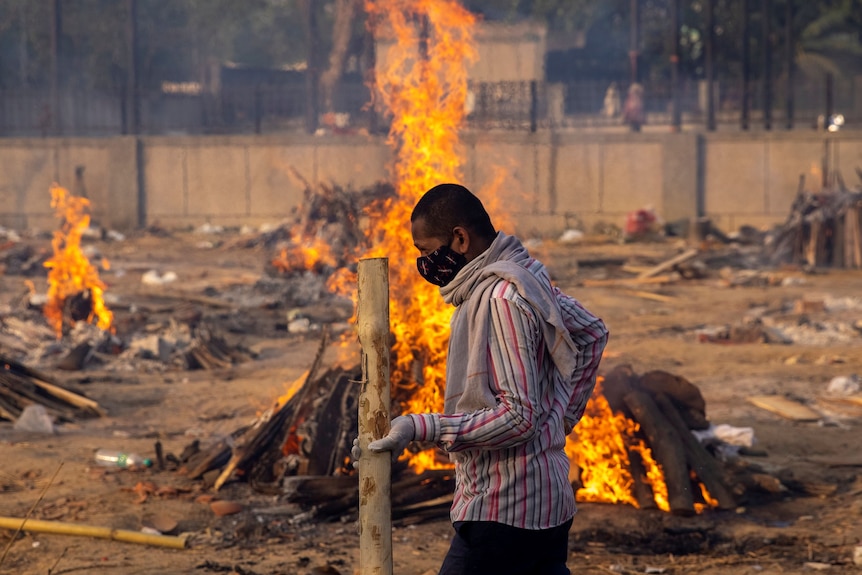 A man walks past burning funeral pyres of people as flames rise behind him.
