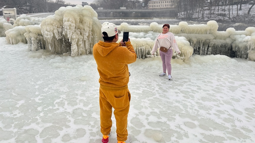 A man in an orange tracksuit takes a picture of a woman in front of ice-covered trees by a river