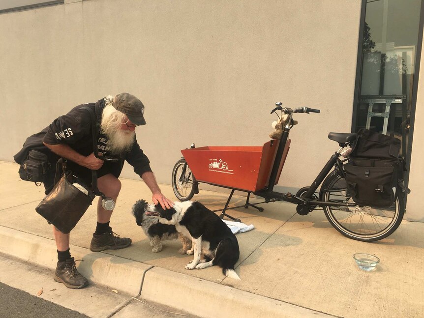 A man with a white beard and hair kneeling down to pat his two dogs.