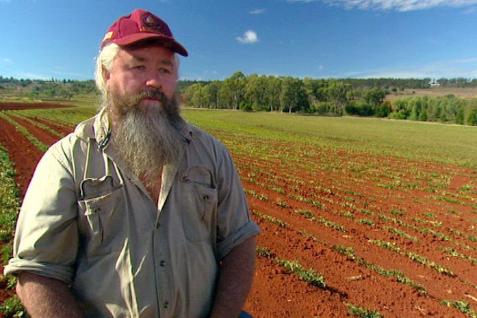 A man with a long beard, wearing a cap and work shirt, stands in a freshly tilled paddock.